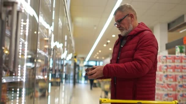 Un homme âgé pensionné avec une barbe grise et des lunettes prend une viande dans un supermarché et la met dans un chariot. Produits laitiers, aliments sains — Video