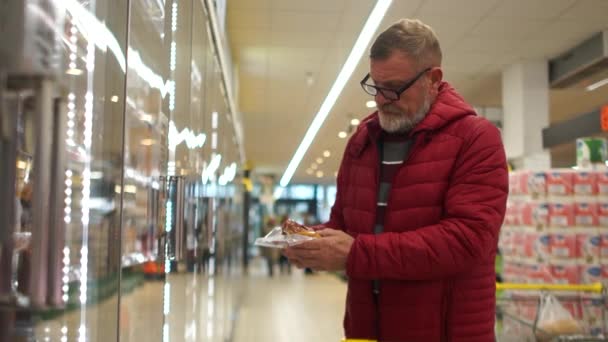 Oudere man gepensioneerde met een grijze baard en een bril pakt een vlees in een supermarkt en zet het in een kar. Zuivel, gezond voedsel — Stockvideo