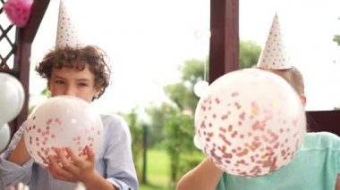 Mutlu çocuklar doğum günü partisi için balon şişirir. Doğum günü şapkalı kız ve erkek balonları pullarla şişirip eğleniyorlar.