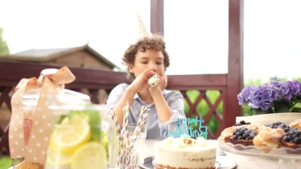 Smutny chłopiec w urodzinowym kapeluszu siedzi przy stole, na którym znajdują się smakołyki i tort z napisem Happy Birthday. Samotne urodziny podczas pandemii covid-19 — Wideo stockowe