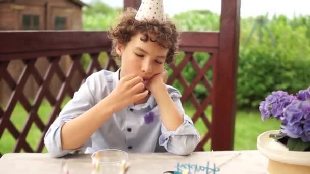 Smutny chłopiec na urodzinach siedzi przy stole, na którym znajdują się smakołyki i tort z napisem Happy Birthday. Samotne urodziny podczas pandemii covid-19 — Wideo stockowe