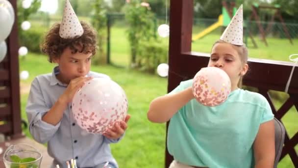 Los adolescentes en sombreros de cumpleaños oy y chica en la fiesta de cumpleaños se divierten rellenando globos grandes con destellos en el interior. Feliz infancia. — Vídeo de stock