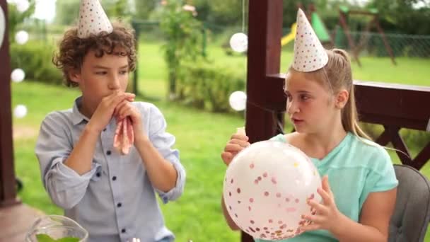 Feliz infancia. Adolescentes en sombreros de cumpleaños oy y chica en la fiesta de cumpleaños divertirse relleno de globos grandes con chispas dentro — Vídeo de stock