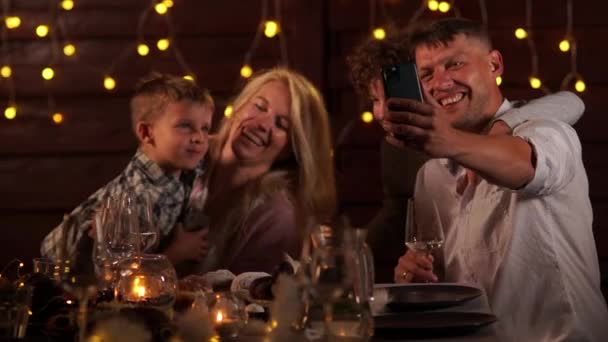 Padre, madre, dos hijos y perro celebran la Navidad en la mesa sobre el fondo de las luces. Videollamada utilizando un teléfono inteligente, distancia social, vídeo felicitaciones — Vídeos de Stock
