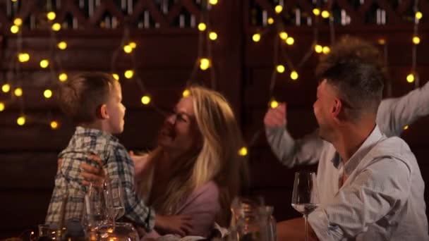 Linda família com dois filhos abraçando enquanto sentado na mesa de Natal no fundo de luzes festivas — Vídeo de Stock
