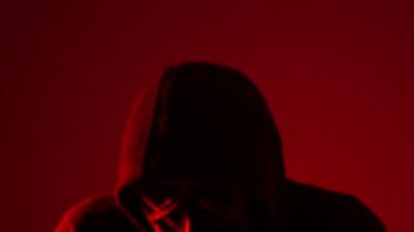 Kırmızı arka planda, neon ışıklı maskeli bir adam. Anonymous kafasını sallıyor, cadılar bayramı maskesi, bilinmeyen korku filmi karakteri, maskeli kötü adam