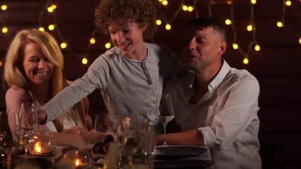 Szczęśliwa rodzina świętuje przyjęcie świąteczne przy stole na tle świateł. perwersyjny nastolatek syn bierze a ciasto z w stół — Wideo stockowe