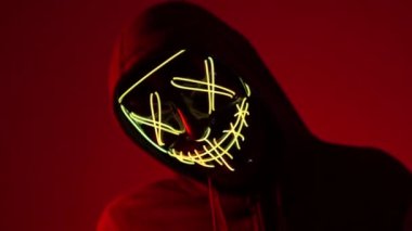 Anonymous kafasını sallıyor, cadılar bayramı maskesi, bilinmeyen korku filmi karakteri, maskeli kötü adam. Kırmızı arka planda, neon ışıklı maskeli bir adam. Kırmızı arka planda neon ışıklı bir adam.