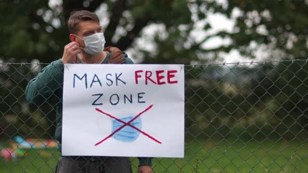 Plakat na ogrodzeniu z napisem Mask Free Zone. Młody aktywista podchodzi i zdejmuje maskę. Powiesiłem go na płocie obok plakatu. Koncepcja anty-maskowa, covid-denier — Wideo stockowe