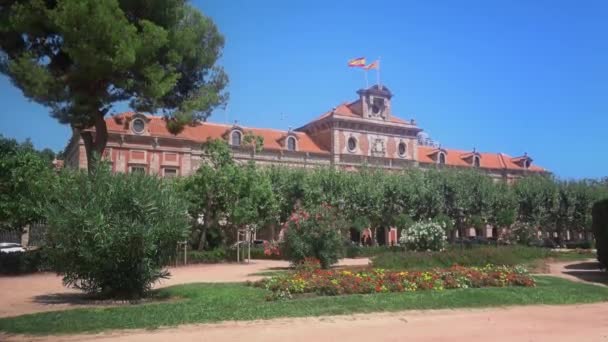 美丽的阳光明媚的街道 花坛和加泰罗尼亚议会背后的树木与西班牙和加泰罗尼亚的旗帜飘扬在建筑物的顶部 — 图库视频影像