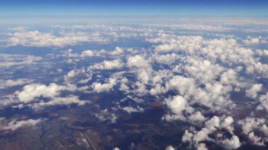 Parlak mavi gökyüzünde sağdan sola yumuşak bir şekilde hareket eden beyaz bulutların güzel yığınları üzerinde uçmak ve kırsal manzaralar çok altında. Uçak penceresinden pitoresk görünüm.