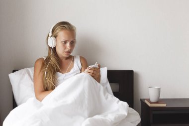 Genç Sarışın ve mutlu gülümseyen kadın müzik dinlemek ve kahve beyaz yatakta sabah zamanında.