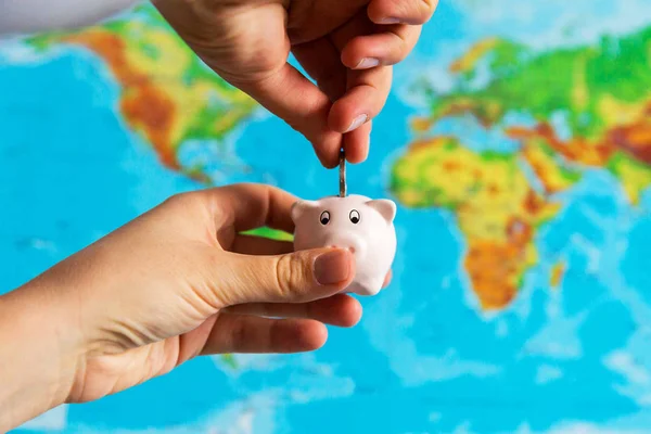 A tiny piggy bank is held in the hand with money. A colorful map of the world in the background. Travel concept.