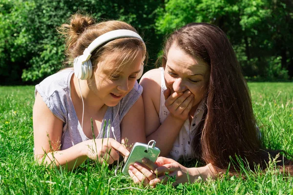 Two beautiful girls: younger and older, read the news in social media on the grass in the city park. Spending time with a friend in the park.