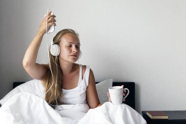 Genç Sarışın ve mutlu gülümseyen kadın müzik dinlemek ve kahve beyaz yatakta sabah zamanında.