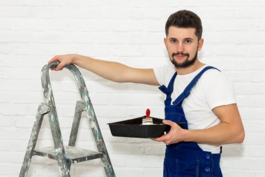 Koyu saçlı Genç adam onun dairesinde beyaz duvar boyuyor. Elinde bir boya pan ve boya fırçası, karşı merdiven yaslanmış tutuyor. Görüntüleyici ve gülümsüyor görünüyor.