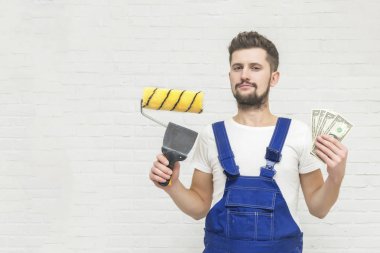 Koyu saçlı Genç adam beyaz daire duvar boyuyor. Elinde bir boya silindiri, kazıyıcı ve para tutar. Ucuz remont kavramı.