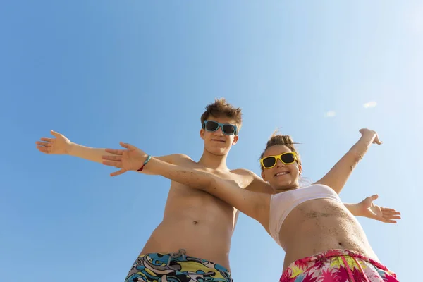 两个年轻美丽的青少年在彩色太阳镜拥抱在海滩上对蓝天 他们靠在相机 看着观众和笑 — 图库照片