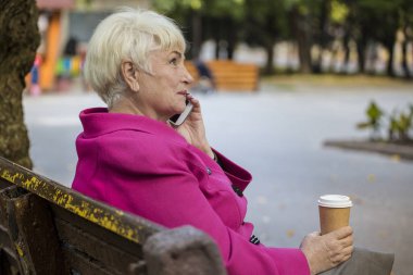 Güzel bir emekli emekli paket servisi olan restoran kahve içer ve bir şehir parkta bir bankta telefonda konuşuyor.