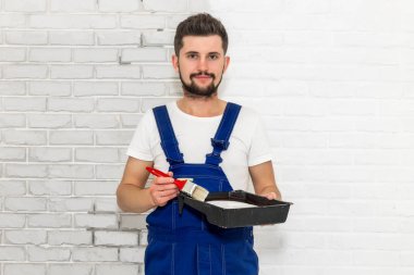 Koyu saçlı Genç adam onun dairesinde beyaz duvar boyuyor. Elinde tutan bir fırça ve boya pan. Görüntüleyici ve gülümsüyor görünüyor.