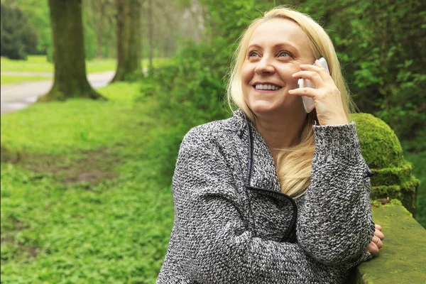 Güzel sarışın kadın 45 yaşında bir cep telefonu üzerinde şehir parkı görüşmelerde gülümseyen.
