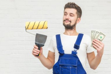 Koyu saçlı Genç adam beyaz daire duvar boyuyor. Elinde bir boya silindiri, kazıyıcı ve para tutar. Ucuz remont kavramı.
