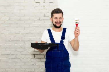 Koyu saçlı Genç adam onun dairesinde beyaz duvar boyuyor. Elinde tutan bir fırça ve boya pan. Görüntüleyici ve gülümsüyor görünüyor.