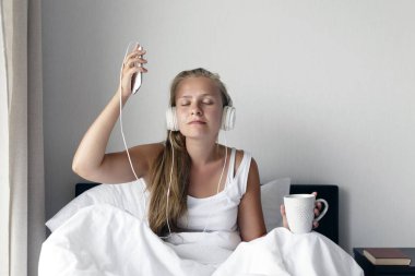 Genç Sarışın ve mutlu gülümseyen kadın müzik dinlemek ve kahve beyaz yatakta sabah zamanında.