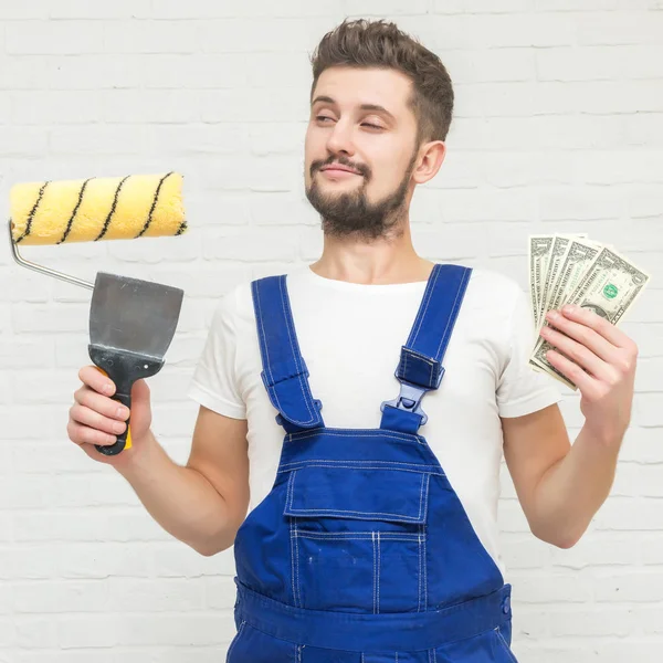 Koyu saçlı Genç adam beyaz daire duvar boyuyor. Elinde bir boya silindiri, kazıyıcı ve para tutar. Ucuz remont kavramı.