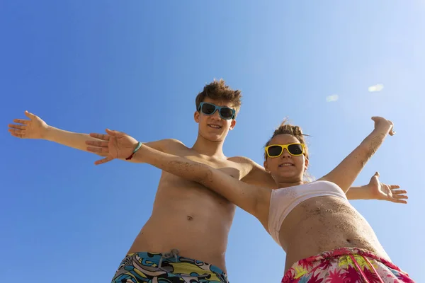 两个年轻美丽的青少年在彩色太阳镜拥抱在海滩上对蓝天 他们靠在相机 看着观众和笑 — 图库照片