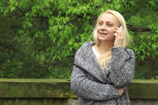 Güzel sarışın kadın 45 yaşında bir cep telefonu üzerinde şehir parkı görüşmelerde gülümseyen.