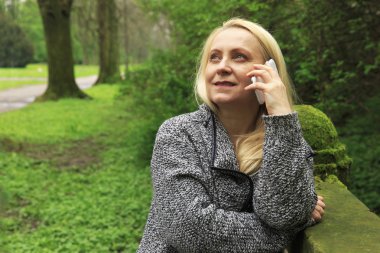 Güzel sarışın kadın 45 yaşında bir cep telefonu üzerinde şehir parkı görüşmelerde gülümseyen.