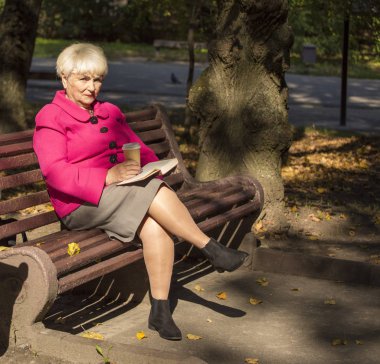 Kentsel bir parkta bir bankta oturmuş bir güzel gülümseyen emekli. O bir kitap ve bir fincan kahve tutar.