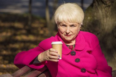 Kentsel bir parkta bir bankta oturmuş bir güzel gülümseyen emekli. O bir kahve onun elinde tutar. Güneşli bir gün. -Kadar yakın.