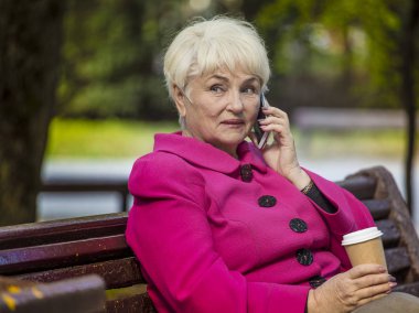 Güzel bir emekli emekli paket servisi olan restoran kahve içer ve bir şehir parkta bir bankta telefonda konuşuyor.