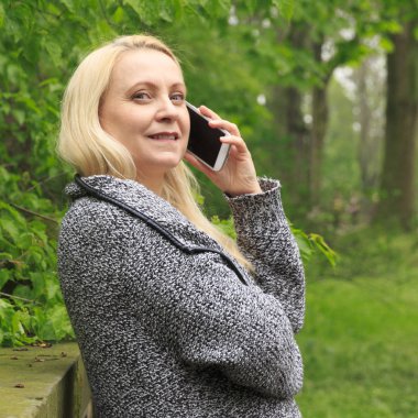 Güzel sarışın kadın 45 yaşında bir cep telefonu üzerinde şehir parkı görüşmelerde gülümseyen.