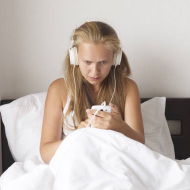 Genç Sarışın ve mutlu gülümseyen kadın müzik dinlemek ve kahve beyaz yatakta sabah zamanında.