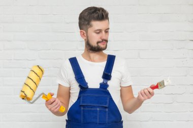 Koyu saçlı Genç adam onun dairesinde beyaz duvar boyuyor. Elinde bir boya silindiri ve boya fırçası tutar. Ne seçmek için karar veremiyorum.