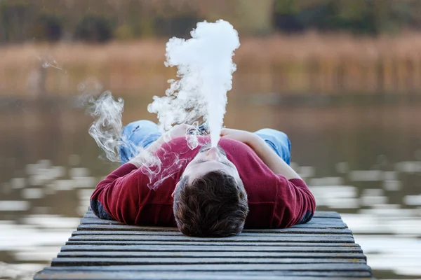 Genç bir adam - bir vaper bir sonbahar gününde bir köprü üzerinde yatıyor ve bir e-sigara içiyor. Arka planda beyaz duman var..