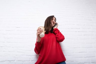 Modern beyaz tuğla loft tarzı duvar arka planı kırmızı kazaklı genç bir çok gülümseyen kız el ve telefon görüşmeleri take-away kahve tutar. Görüntüleyiciyi görünüyor. Kopya alanı.