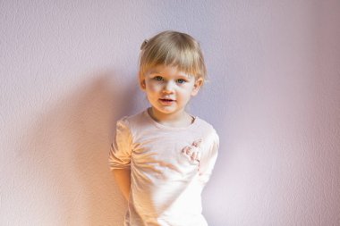 Üzgün küçük kız sarışın saçlı güneşli bir odada evde duvara yaslanmış. Görüntüleyiciyi görünüyor. Görünümü en çok, yakın çekim.