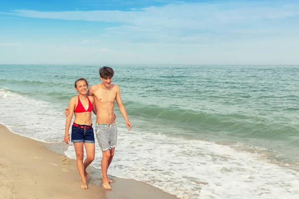 二十代の若者 少女とブロンドの髪を持つ少年が服を水着姿で歩く海のビーチで視聴者へ帰国 — ストック写真