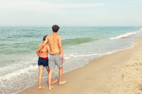 二十代の若者 女の子と水着を着て ブロンドの髪を持つ少年が海のビーチに歩いています 背面図 — ストック写真