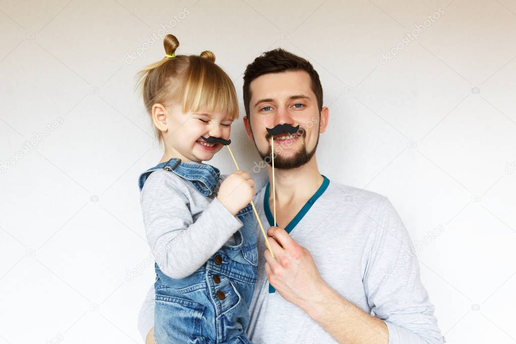 Fathers day! A blonde girl wearing a denim dungarees, hugs and laughs with her dad. They are holding mustaches in their hands. White background, selective focus.