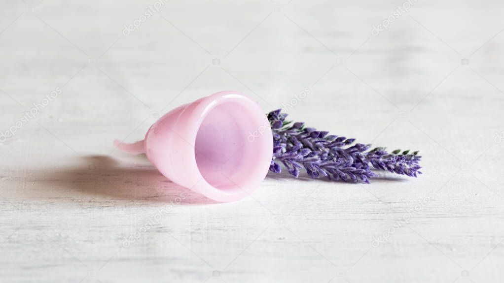 Pink menstrual cup lies on a white vintage wooden background. Next there are lavender flowers. Selective focus, copy space.