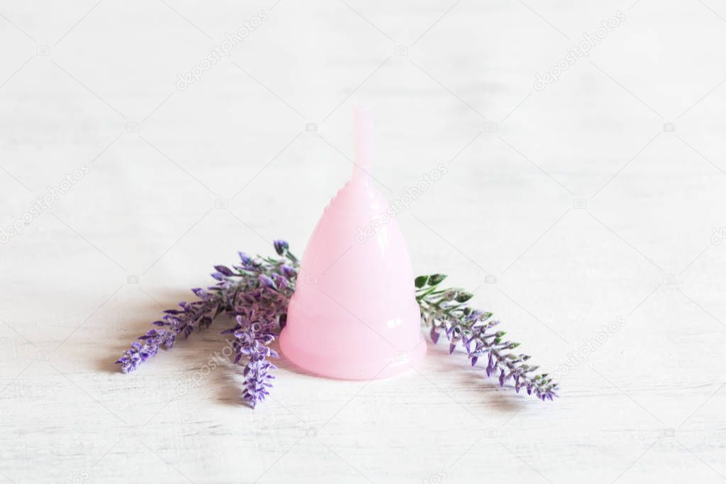 Pink menstrual cup lies on a white vintage wooden background. Next there are lavender flowers. Selective focus, copy space.