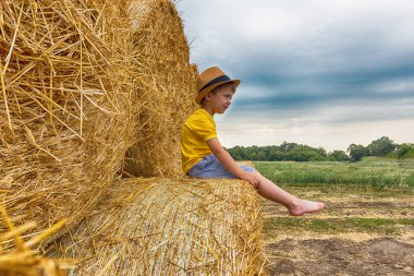 Küçük güzel çocuk bir sarı tişört ve kot şort giymiş ve modern şapka bir haystack bir yaz günü oturur. Hasat.