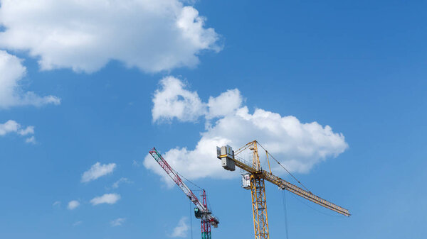 Two tower cranes against a sunny sky with white clouds.