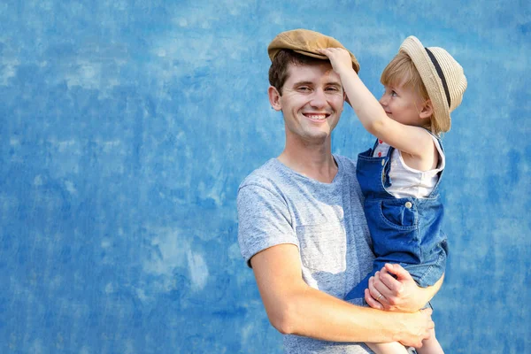 A young dad is holding his daughter on his hands, laugh and have fun on a summer, sunny day. Blue background. Copy space.