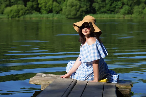 Köprü Nehri yakınında oturan mutlu genç kadın. Mavi bir elbise ve hasır şapka. Arkadan görünüm.